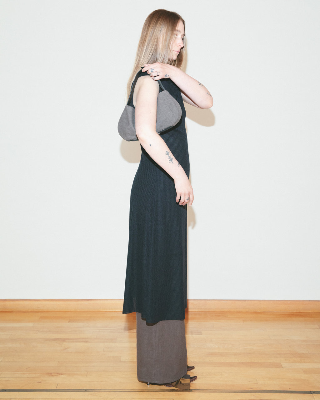 Woman wearing a long black dress with a gray shoulder bag against a white wall.