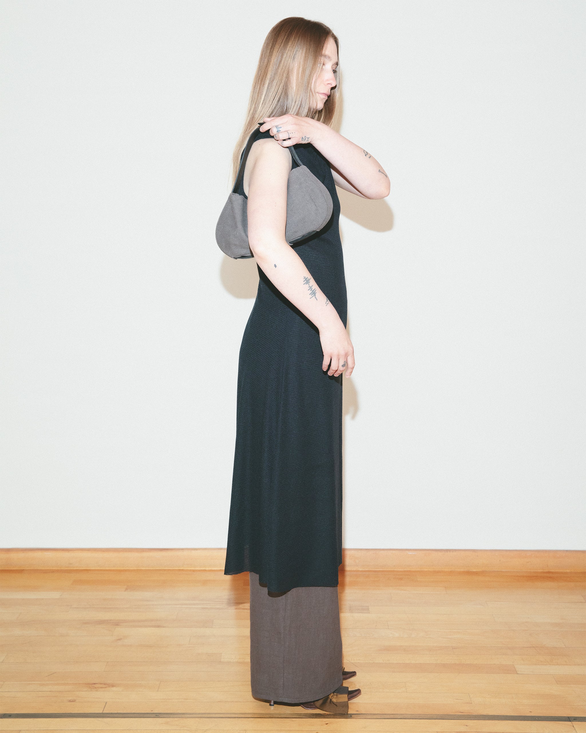 Woman wearing a long black dress with a gray shoulder bag against a white wall.
