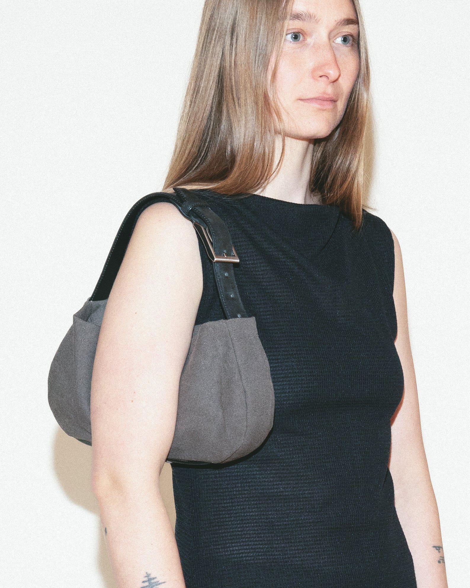 Woman wearing a long black dress with a gray shoulder bag against a white wall.